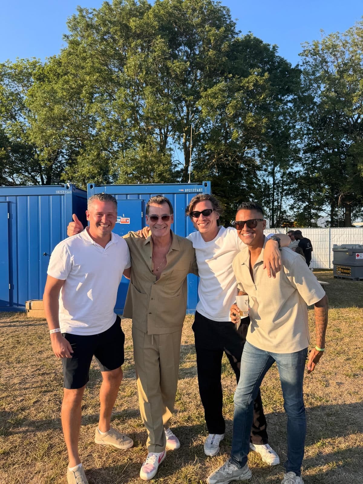 Fergino met drie anderen, lachend en met de armen om elkaar heen, op een evenemententerrein in de avondzon