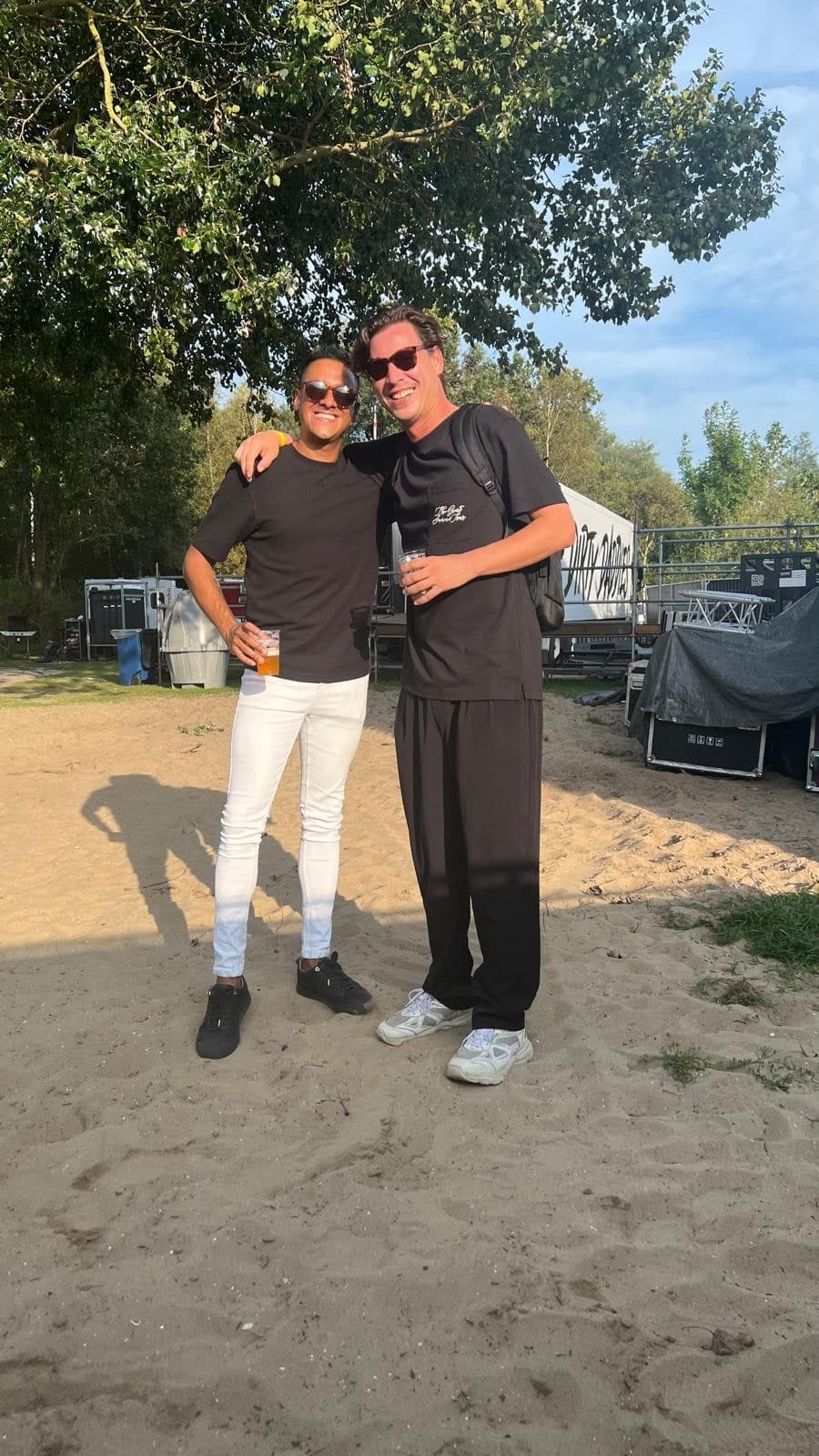Tony en Fergino, lachend en met de armen om elkaar heen, op een zandstrand bij een evenemententerrein in de avondzon